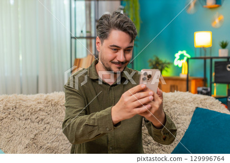 Middle-aged man happy smiling nodding reading smartphone message relaxing on home sofa couch indoors Middle-aged man happy smiling nodding reading smartphone message relaxing on home sofa couch indoors 129996764