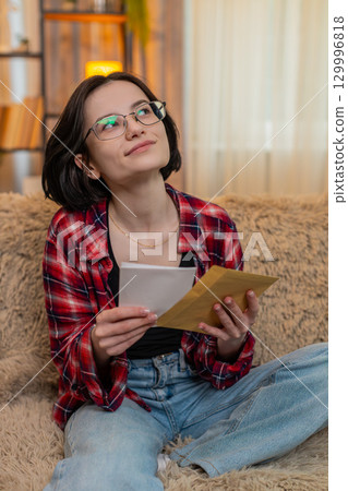 Woman writing heartfelt letter on paper at home, smiling with emotion before sealing it in envelope 129996818