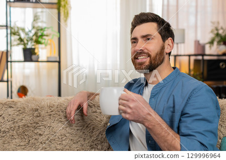 Young man sitting on sofa holding a cup drinking tea, gazing into distance enjoying the moment Young man sitting on sofa holding a cup drinking tea, gazing into distance enjoying the moment 129996964