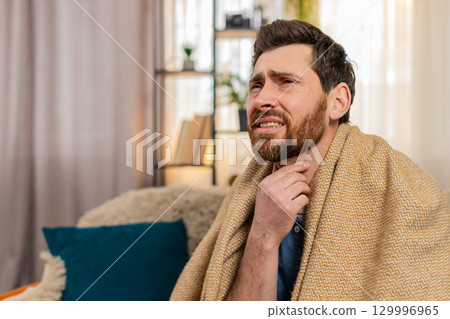 Young man sitting on the sofa at home, wrapped in a blanket, coughing, holding throat, feeling pain 129996965