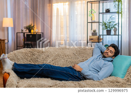 Tired Indian man lying on comfortable couch taking rest and daytime nap in living room at home 129996975