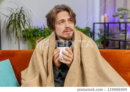 Sick ill Caucasian man wrapped in plaid sit shivering from cold drinking hot coffee or tea at home 129996999