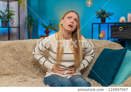 Caucasian woman abdominal stomach pain on sofa holding belly cramps suffering indoors discomfort 129997007