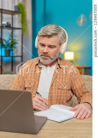 Mature man with laptop and headphones, listening to podcast making notes education study at home 129997009