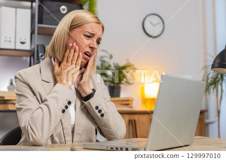 Annoyed mature businesswoman looking at laptop screen irritated working remotely at office home 129997010