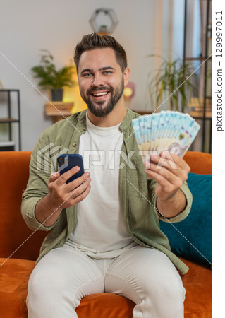 Successful rich happy young winner man holding Ukrainian hryvnias UAH money bill fan on home sofa Successful rich happy young winner man holding Ukrainian hryvnias UAH money bill fan on home sofa 129997011
