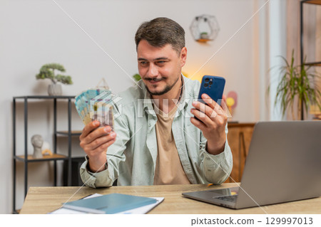 Smiling Caucasian young man counting money cash using smartphone at home office table in living room Smiling Caucasian young man counting money cash using smartphone at home office table in living room 129997013