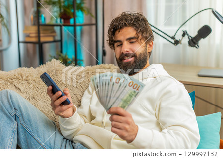 Successful rich young Indian man holding dollar bill and smartphone sitting on sofa couch at home Successful rich young Indian man holding dollar bill and smartphone sitting on sofa couch at home 129997132