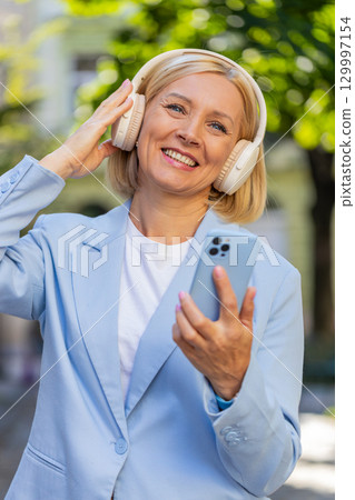 Happy carefree mature businesswoman with smartphone listening to music in headphones on city street 129997154