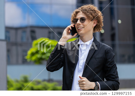 Businessman making phone call online conversation loudspeaker talking waving hello outside office Businessman making phone call online conversation loudspeaker talking waving hello outside office 129997155