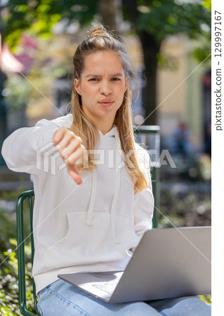 Upset young woman lady using laptop looking at camera and showing thumb down in city street outdoors Upset young woman lady using laptop looking at camera and showing thumb down in city street outdoors 129997167