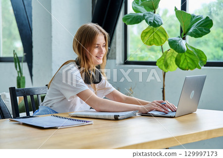 Young teenager girl college student sitting at desk with laptop, typing writing studying 129997173