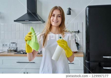 Smiling young woman in gloves with rag cleaning spray looking at camera in kitchen Smiling young woman in gloves with rag cleaning spray looking at camera in kitchen 129997177