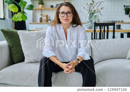 Successful beautiful mature woman looking at camera sitting at home on sofa 129997188
