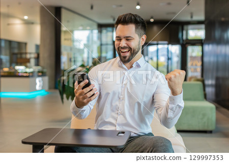 Happy young businessman using smartphone shocked by sudden victory lottery win in office lobby Happy young businessman using smartphone shocked by sudden victory lottery win in office lobby 129997353