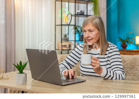Mature old woman working from home on laptop, writing report, replying emails, sipping coffee Mature old woman working from home on laptop, writing report, replying emails, sipping coffee 129997361