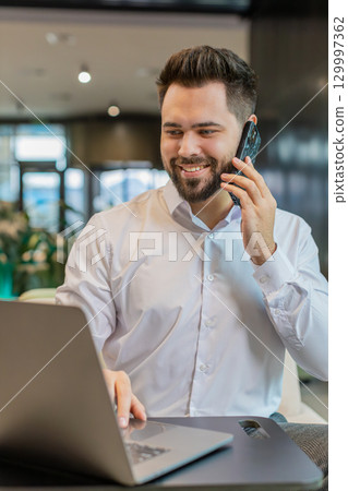 Businessman in formal shirt answering to client on mobile phone call, using laptop in office lobby Businessman in formal shirt answering to client on mobile phone call, using laptop in office lobby 129997362