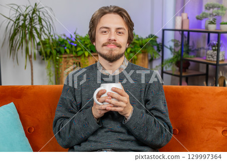 Handsome smiling Caucasian young man drinking a cup of coffee or herbal tea sitting at home couch Handsome smiling Caucasian young man drinking a cup of coffee or herbal tea sitting at home couch 129997364