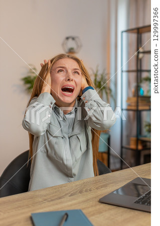 Frustrated freelancer using laptop struggling to work due to loud noise sitting at home office table Frustrated freelancer using laptop struggling to work due to loud noise sitting at home office table 129997366
