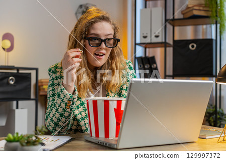 Businesswoman taking break from work wearing 3D glasses eating popcorn and watching movie at office Businesswoman taking break from work wearing 3D glasses eating popcorn and watching movie at office 129997372