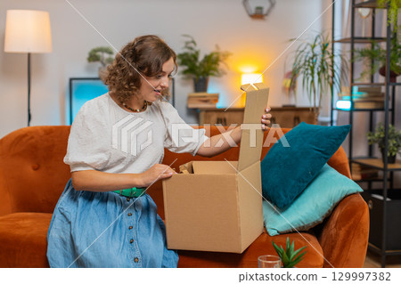 Smiling satisfied young woman shopper online shop customer opening unpacking cardboard box on sofa 129997382