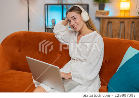 Happy overjoyed young woman girl in wireless headphones listening music sitting on sofa at home 129997392
