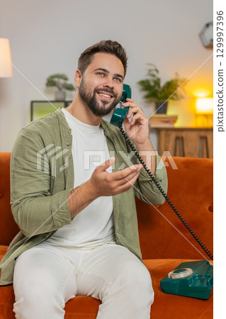 Smiling young man making wired telephone call conversation with friends sitting on sofa at home 129997396