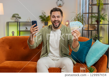 Successful rich happy young winner man holding Ukrainian hryvnias UAH money bill fan on home sofa 129997434