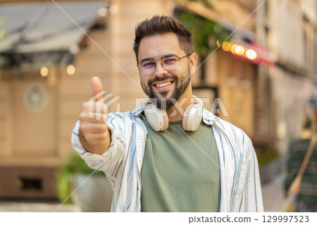 Cheerful young Caucasian man showing double thumbs up gives positive reply standing on city street 129997523