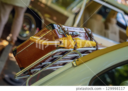 Suitcase and umbrella in the trunk. Close-up of vintage suitcase and yellow umbrella strapped on retro car’s luggage rack. Concept of nostalgic road trip, summer getaway, travel aesthetic Suitcase and umbrella in the trunk. Close-up of vintage suitcase and yellow umbrella strapped on retro car’s luggage rack. Concept of nostalgic road trip, summer getaway, travel aesthetic 129998117