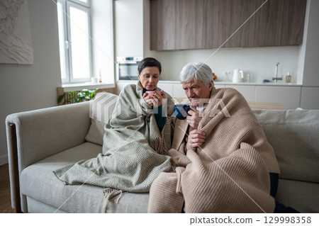 Upset struggling senior couple under blanket sipping hot tea remedy, suffer from illness at home 129998358