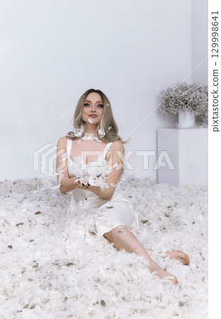 a very beautiful blonde sits in a white room in which feathers are flying 129998641