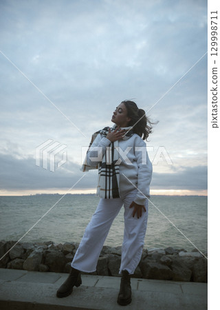 Beautiful young girl in a white tracksuit stands on the seashore in stormy weather 129998711