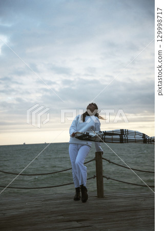 Beautiful young girl in a white tracksuit stands on the seashore in stormy weather 129998717