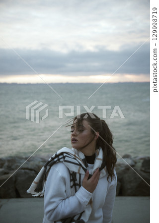 Beautiful young girl in a white tracksuit stands on the seashore in stormy weather Beautiful young girl in a white tracksuit stands on the seashore in stormy weather 129998719