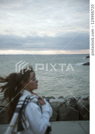 Beautiful young girl in a white tracksuit stands on the seashore in stormy weather Beautiful young girl in a white tracksuit stands on the seashore in stormy weather 129998720