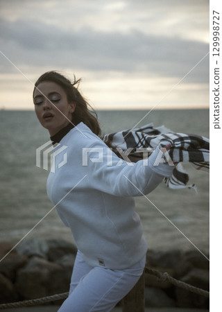 Beautiful young girl in a white tracksuit stands on the seashore in stormy weather 129998727
