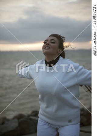 Beautiful young girl in a white tracksuit stands on the seashore in stormy weather 129998728