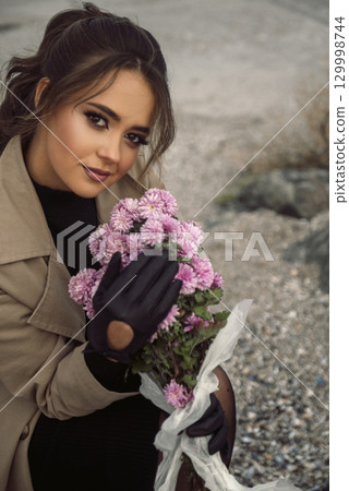 brunette girl in beige coat sits on chair among rocks 129998744
