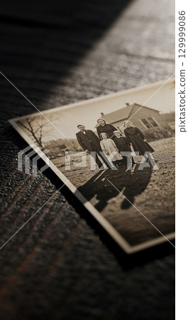 A sepia-toned print of a five-person family, including a man, woman, and three children, standing outdoors in front of a modest house, resting on a dark, textured wooden surface with dramatic. 129999086