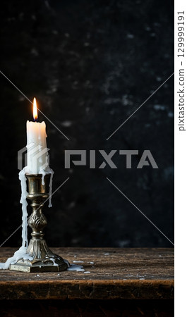 An elegant white candle, heavily dripped with wax, stands in an antique silver candlestick on a rough wooden surface, casting a warm glow against a dark wall. 129999191