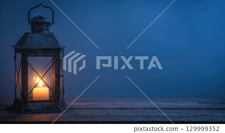An old, rustic metal lantern with frosted glass panes and a lit candle inside sits on a wooden surface against a dark blue background, offering warm light and copy space. 129999352