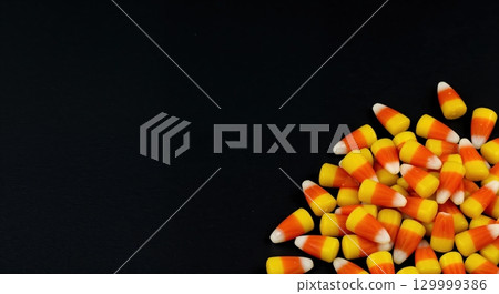 A vibrant pile of classic candy corn with distinct yellow, orange, and white stripes rests neatly in the bottom right corner against a dark, plain black background. 129999386