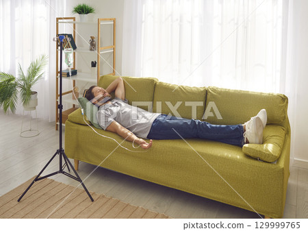 Smiling man lying on the couch at home while receiving IV drip infusion and vitamin therapy. Smiling man lying on the couch at home while receiving IV drip infusion and vitamin therapy. 129999765