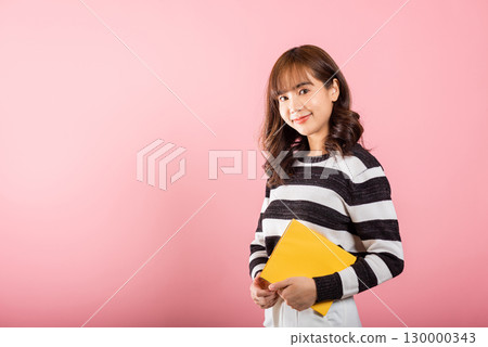 Portrait of beautiful Asian young woman teenage smiling holding a yellow book and reading, studio shot isolated on pink background with copy space 130000343