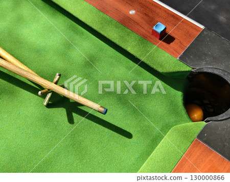 Billiard table with cue stick, bridge, chalk, and ball near the pocket under sunlight. Billiard table with cue stick, bridge, chalk, and ball near the pocket under sunlight. 130000866