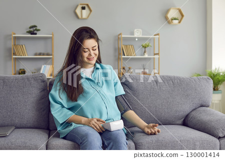 Happy young pregnant woman sitting on the sofa at home and measuring blood pressure. 130001414