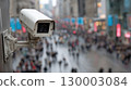Overhead white security camera observing a bustling urban street. Blurred crowds move along the wet outdoor public space. Electronic eye monitors daily city activity and safety. 130003084