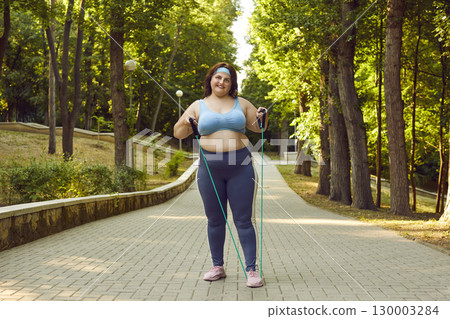 Fat woman doing sport exercises with rubber band standing in summer park outdoors. Fat woman doing sport exercises with rubber band standing in summer park outdoors. 130003284