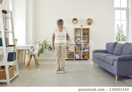 Senior woman standing on weight scales in the living room and looking down at figures Senior woman standing on weight scales in the living room and looking down at figures 130003340